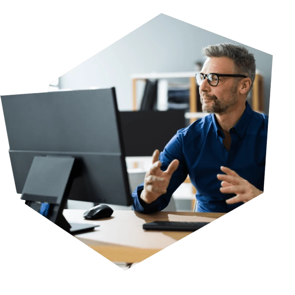 Mann im Business Outfit sitzt vor seinem Schreibtisch auf dem ein großer Monitor steht und nimmt an einem Online-Meeting über Microsoft Teams teil