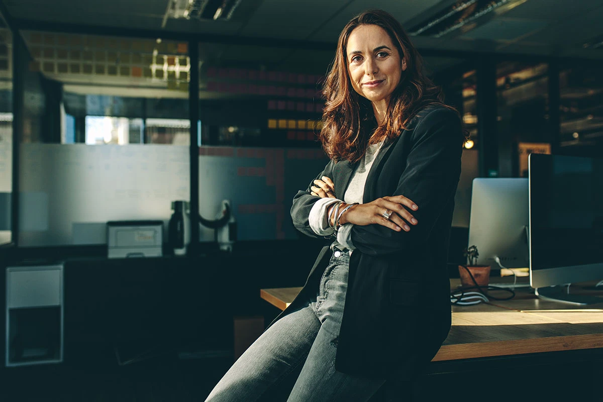 weibliche F&uuml;hrungskraft schaut mit verschr&auml;nkten Armen und anlehnend an einen Schreibtisch in die Kamera in einem modernen Sonnenlicht geflutetet B&uuml;ro