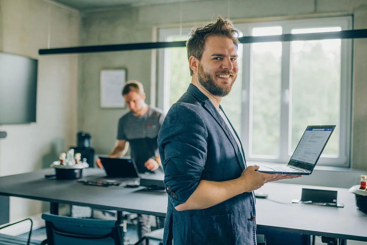 Mitarbeiter mit Laptop in der Hand schaut seitlich in die Kamera, im Hintergrund ist ein moderner Meetingraum zu sehen an dem ein weiterer Mitarbeiter an seinem Laptop steht