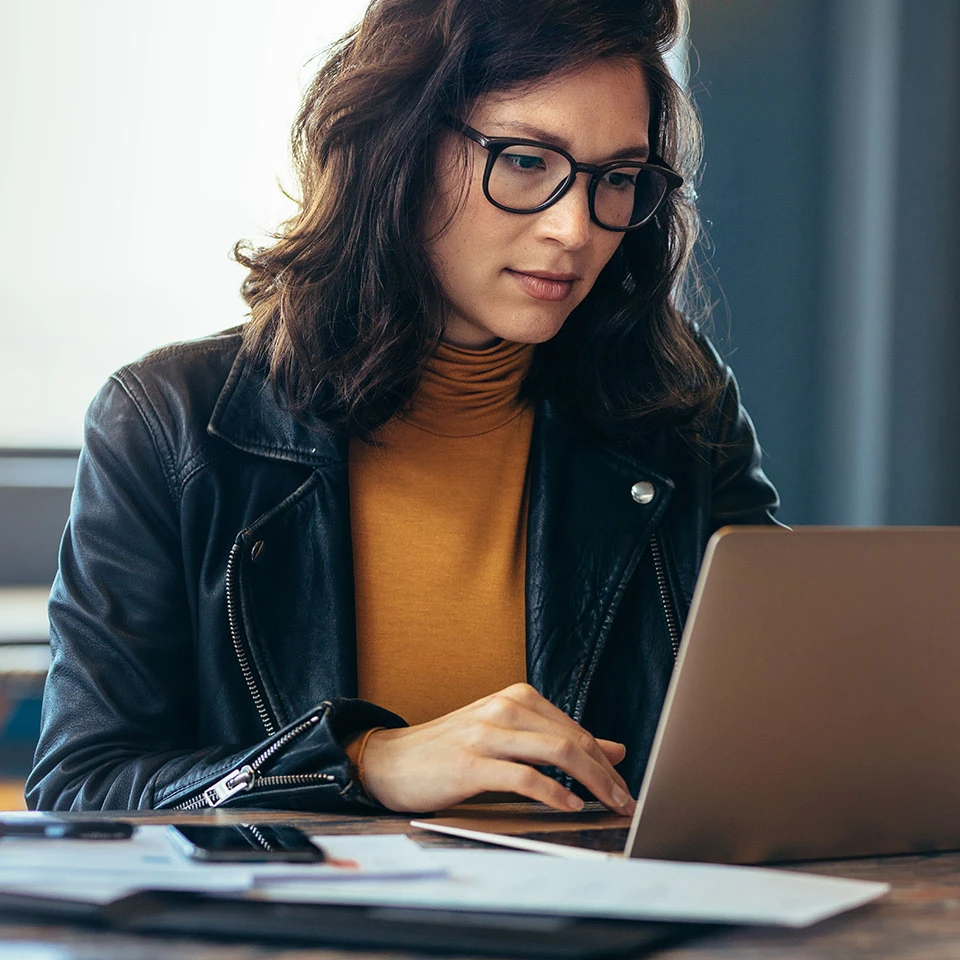 Frau arbeitet an einem Schreibtisch an einem Laptop, sie trägt eine schwarze Lederjacke sowie einen orangenen Rollkragenpullover