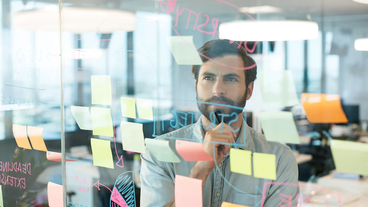 Fotografiert durch eine Glasscheibe auf deren Seite ein Mann auf viele farbige Post-Its schaut, die auf der Scheibe kleben, und das Wort Strategie drauf geschrieben ist