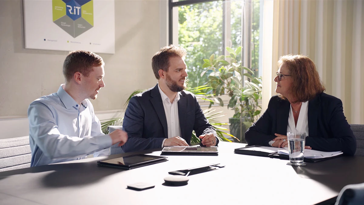 zwei M&auml;nner und eine Frau sitzen in einem modernen B&uuml;ro, vor ihnen liegen Tablets und Arbeitsmaterialien