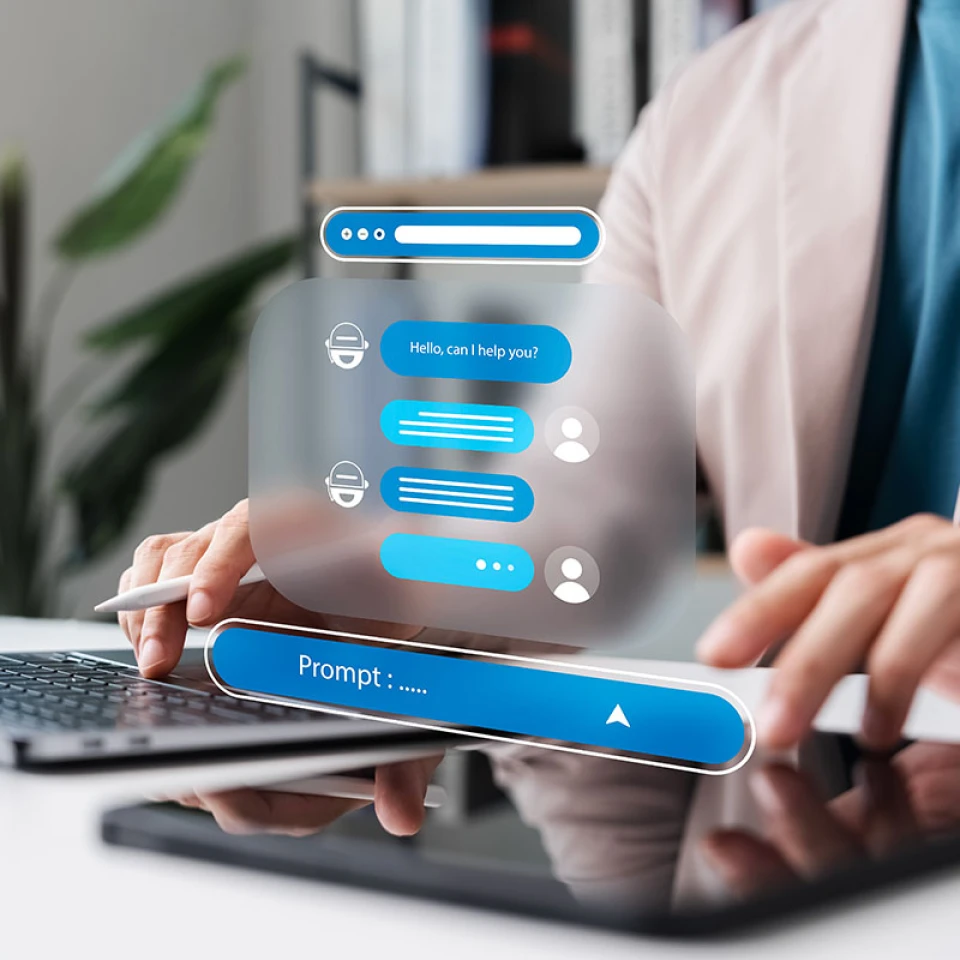 Person arbeitet vor ihrem Laptop und mit einem Tablet, ein grafisches Element schwebt über dem Tablet und zeigt den Chatverlauf mit einem Chatbot