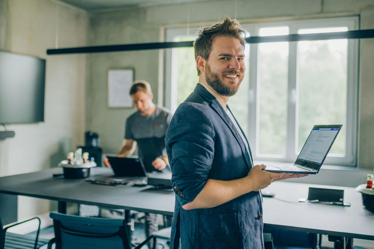 Mann im Business Outfit hält seinen Laptop vor sich und schaut in die Kamera im HIntergrund ist ein moderner Konferenzraum zu sehen an dem ein weiterer Mann arbeitet