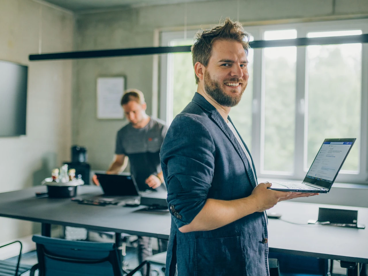Mann im Business Outfit hält seinen Laptop vor sich und schaut in die Kamera im HIntergrund ist ein moderner Konferenzraum zu sehen an dem ein weiterer Mann arbeitet