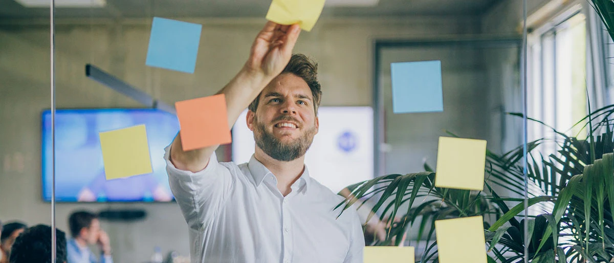 Mann im weißen Hemd klebt farbige Post-Its an eine Glasscheibe im Hintergrund ist ein moderner Konferenzraum mit Teilnehmern zu sehen