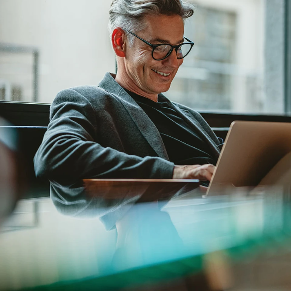 Mann mit grauen Haaren und modernen Business Outfit sitzt auf einer Couch und arbeitet an einem Laptop